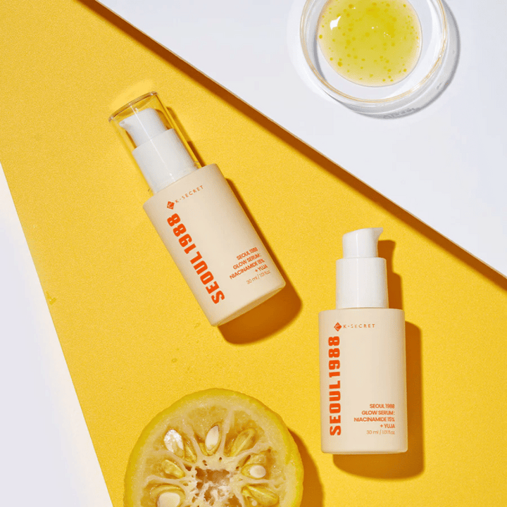 Två flaskor Seoul 1988 Glow Serum placerade på gul bakgrund tillsammans med en skål med gult serum och en halverad citrusfrukt.