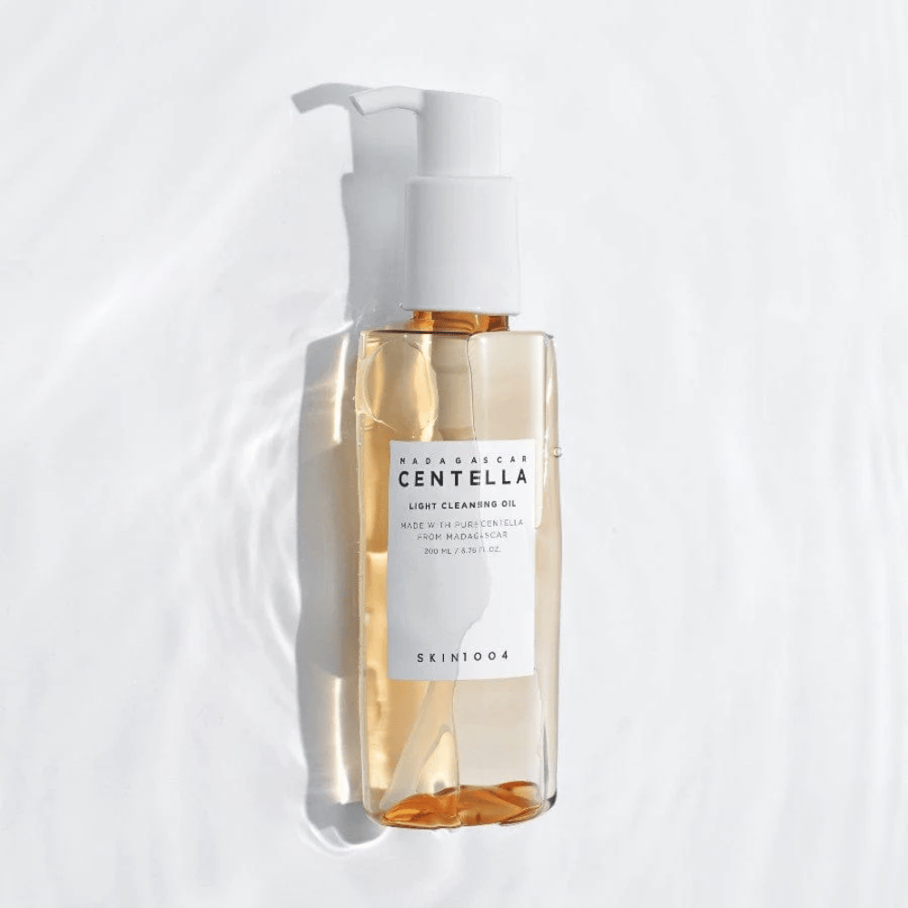 SKIN1004 Madagascar Centella Light Cleansing Oil på vit bakgrund med mjuka vattenreflexer som framhäver den transparenta flaskan och gyllene innehållet.