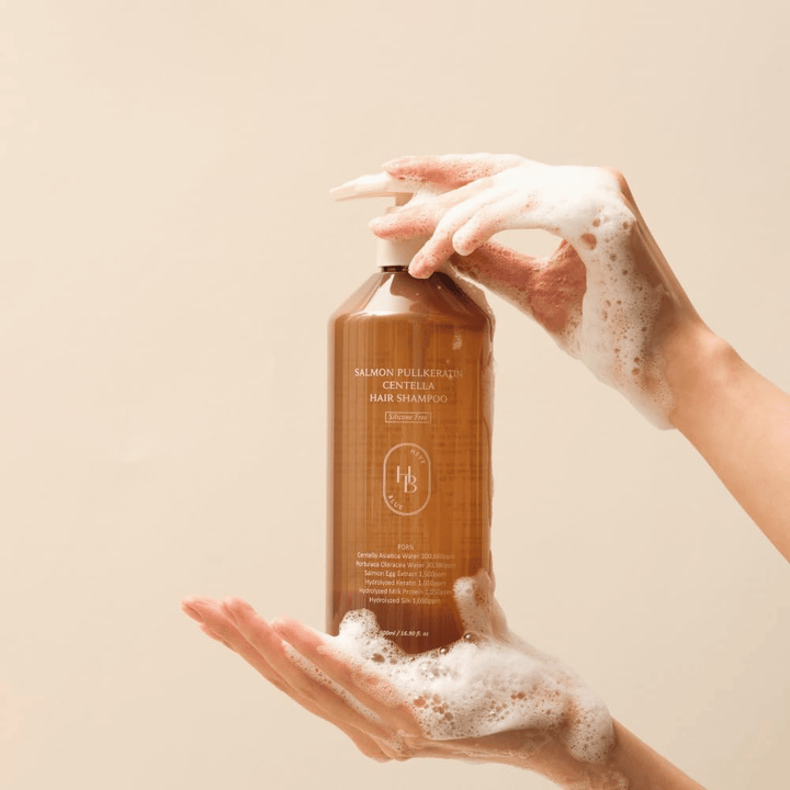 Heveblue Salmon Pullkeratin Centella Hair Shampoo in a brown bottle surrounded by foam. The product is held between two hands and highlights a cleansing and nourishing hair care experience with abundant lather.