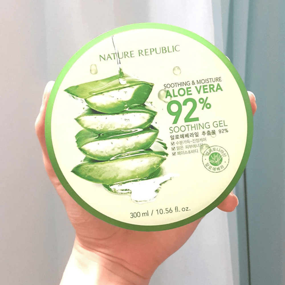 A hand holds up a round jar with Nature Republic Aloe Vera 92% Soothing Gel. The jar is green with an image of aloe vera and text describing its soothing and moisturizing properties. The content is 300 ml.