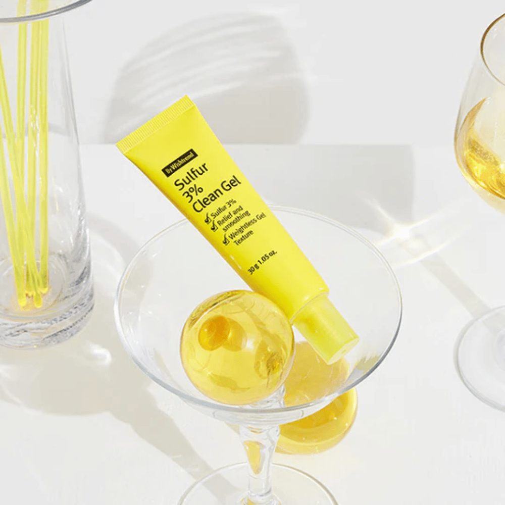 By Wishtrend Sulfur 3% Clean Gel in a yellow tube placed in a cocktail glass with a yellow sphere, surrounded by elegant glasses on a white background.