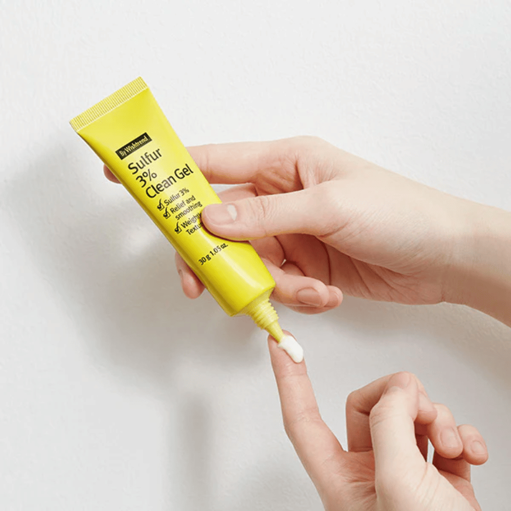Close-up of hands applying By Wishtrend Sulfur 3% Clean Gel, a yellow tube with white gel, against a white background.