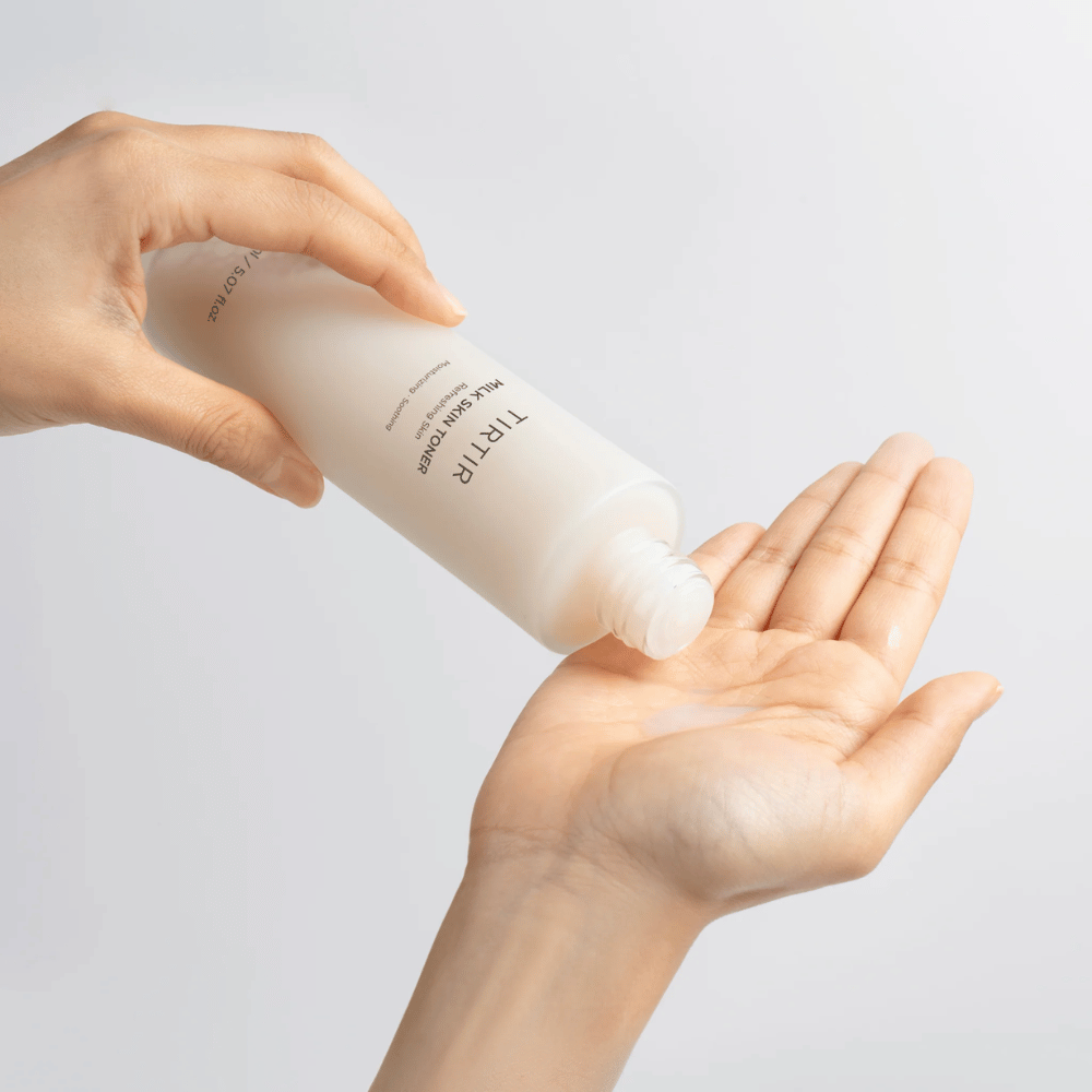 The image shows a person pouring a milky liquid from a bottle labeled "TIRTIR Milk Skin" onto the palm. The product seems to be a skin toner or moisturizing lotion.