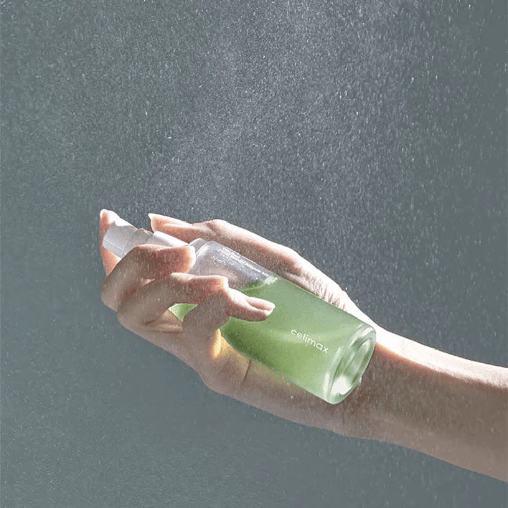 A hand holds Celimax Noni Mist spray bottle and applies the product in a fine, even spray on the skin. The bottle is green and the product is marketed as "The Real Noni Energy Ampule Mist" to provide energy and hydration to the skin.