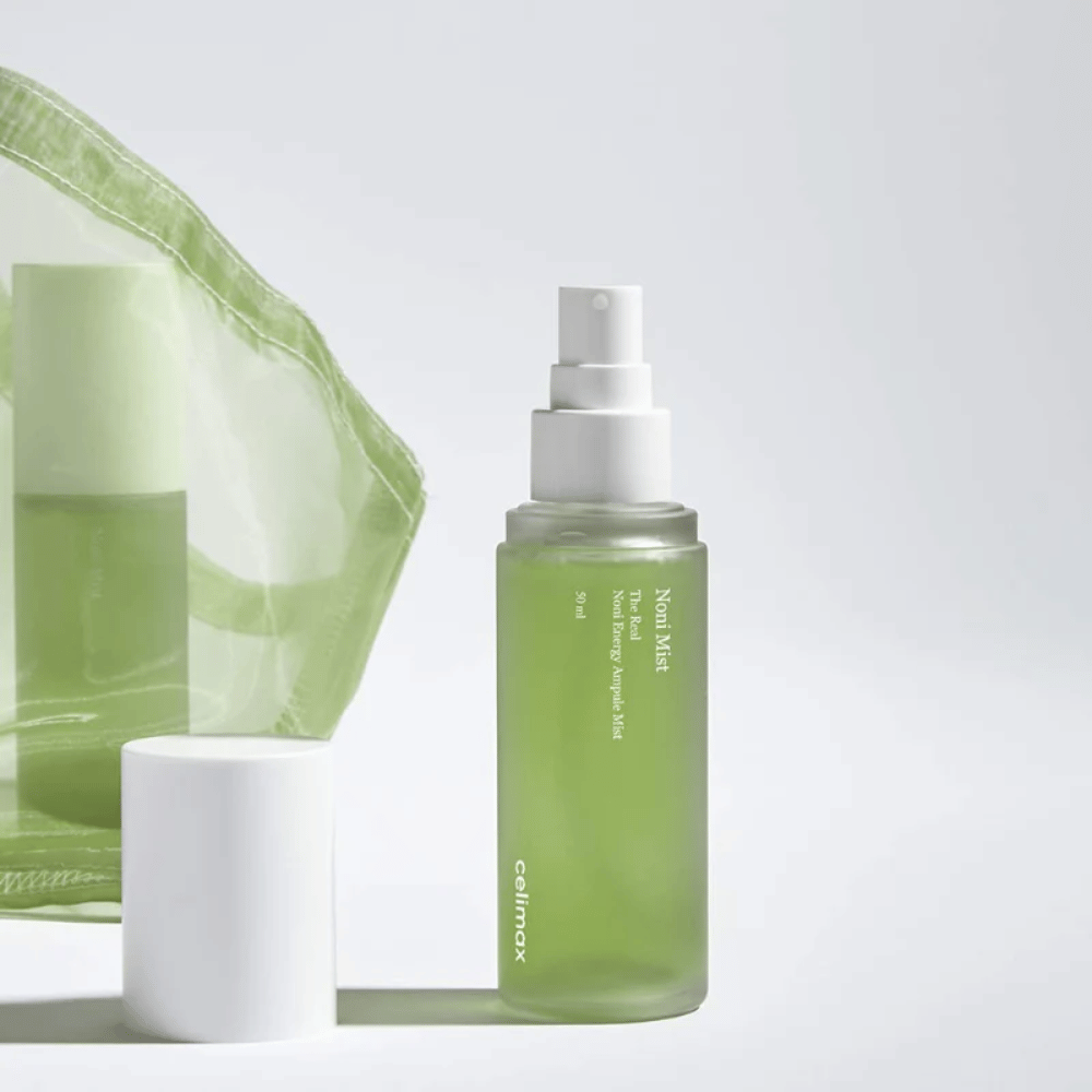 A bottle of Celimax Noni Mist, a green spray bottle with a white pump, standing next to a cap. The product is packaged in a transparent plastic bag and marketed as Noni Energy Ampule Mist.