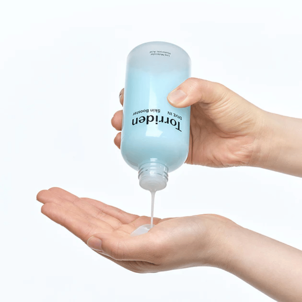 Hands holding a bottle of skincare product and dispensing a clear liquid into the palm. The bottle is labeled with "Torriden - DIVE-IN Low Molecule Hyaluronic Acid Skin Booster".