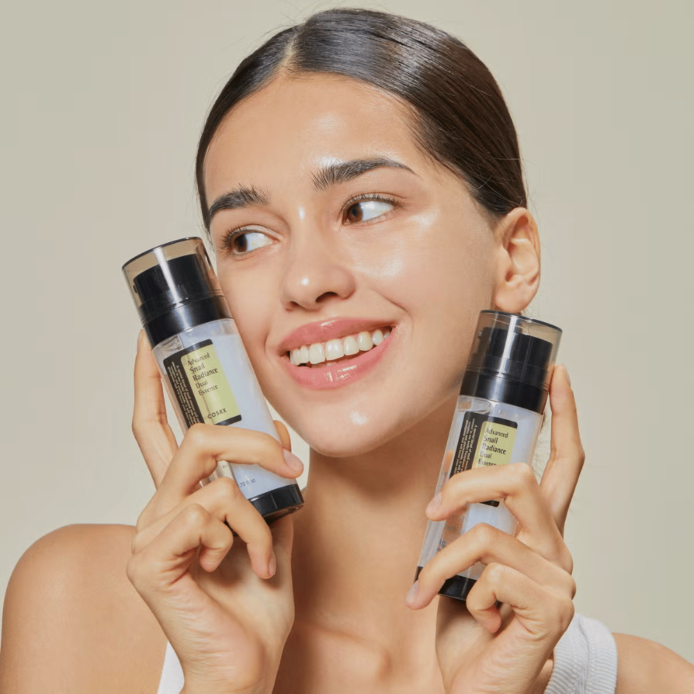 Smiling woman with glossy skin holds two bottles COSRX Advanced Snail Radiance Dual Essence 80 ml near her face against a light background.