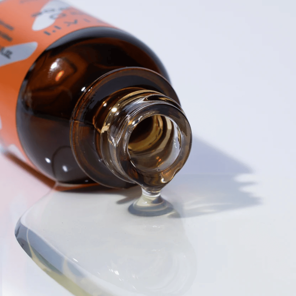 Close-up of TIA'M Vita C Source serum pouring from a brown glass bottle. The serum's clear formula highlights its concentrated vitamin C content for radiant skin.