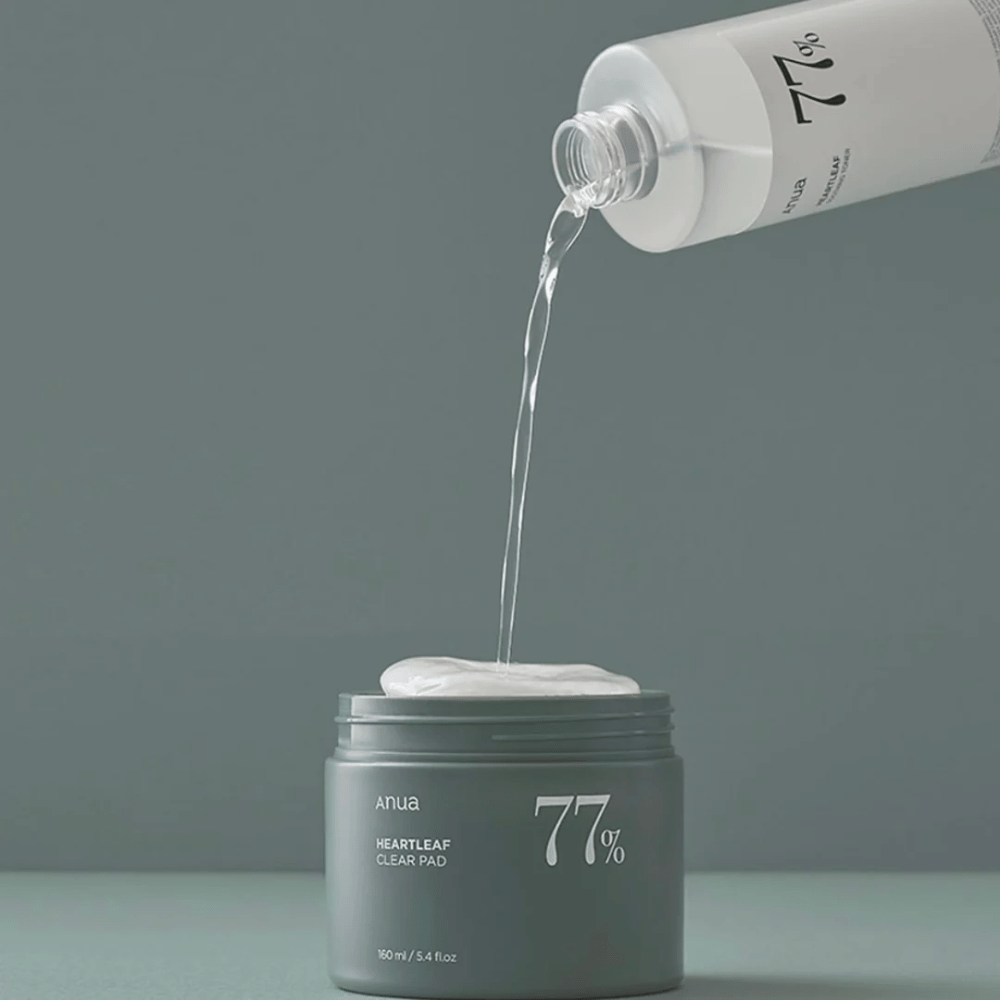 A transparent bottle pours a liquid into a gray jar labeled "anua HEARTLEAF 77% Clear Pad", which contains a stack of white skincare pads. The background is simple and solid-colored in a soft gray tone.